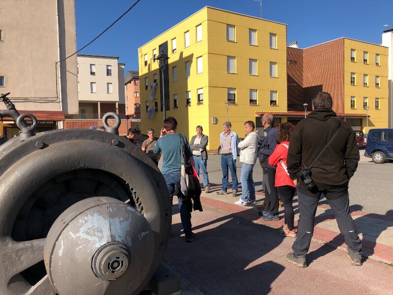 Grupo de personas en corro delante de unos edificios y junto a una turbina eléctrica