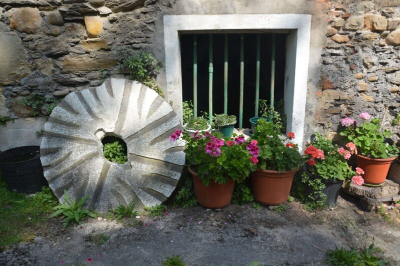 Rueda de piedra para moler cereal apoyada en una pared de piedra. Unas macetas con flores decoran la imagen.